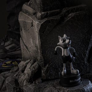 May include: A cartoon raccoon figurine with a black and white color scheme. The figurine is standing on a black base that reads "Mr. Raccoon". The raccoon has black gloves and shorts. The background is a dark, rocky environment.