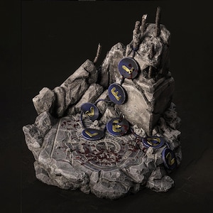 May include: A detailed miniature diorama featuring a stone structure with a circular base and jagged edges. Dark blue circular charms with gold lettering are connected by a blue string. The base has a pattern and red splatters.
