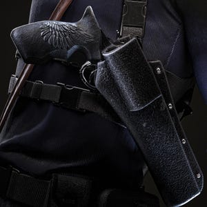 May include: Close-up of a black pistol with an ornate wing design on the handle, holstered on a person's side. The holster is textured and secured with a belt and straps. The person is wearing a dark blue uniform.