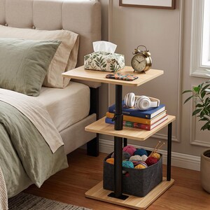 May include: A three-tiered side table with a light wood grain finish and black metal supports. The top shelf holds a tissue box, an alarm clock, and a phone. The middle shelf holds books and headphones. The bottom shelf holds a felt basket of yarn.