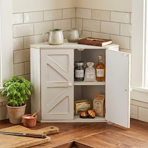 May include: A white corner cabinet with two doors featuring a diagonal panel design. The cabinet interior holds a glass jar of Earl Grey tea, a bag of Fleur de Sel, a bottle of artisan balsamic glaze, a box of cultured butter, and a bag of coffee.