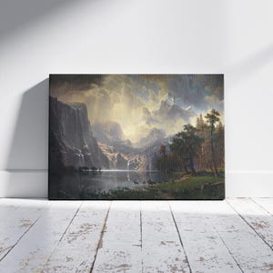 Pode incluir: Uma pintura de paisagem emoldurada retratando um vale montanhoso com um lago, árvores e vida selvagem. A obra de arte apresenta um céu dramático com luz rompendo as nuvens, iluminando a cena. A pintura é exibida em uma parede branca e repousa sobre um chão de madeira branca.