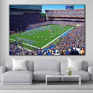 May include: A large canvas print of a football stadium filled with spectators. The field is green with white yard lines and the word "Florida" in blue. The stands are packed with fans wearing blue and orange. A marching band is on the field.