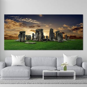 May include: A large rectangular canvas print depicting Stonehenge at sunset. The ancient stone structure stands on a green field under a dramatic sky of orange and blue hues. The artwork is displayed above a gray sofa.
