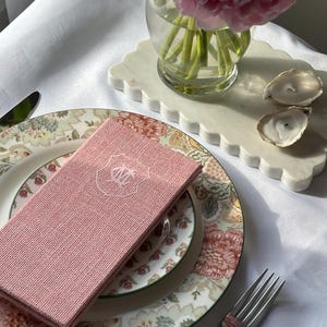 May include: A rectangular, textured, dusty pink linen napkin featuring a white embroidered crest with the letters DM in the center. The napkin is folded and placed on a floral patterned ceramic plate with a silver fork resting nearby.