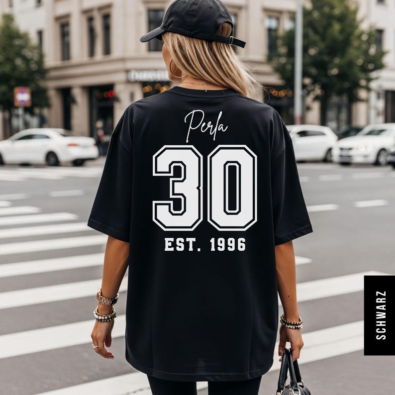 Op de afbeelding: Zwart oversized T-shirt met het woord "Perla" in script boven het nummer "30" in een vetgedrukte, omlijnde letter. Daaronder staat "EST. 1996". De persoon draagt het shirt op een zebrapad.