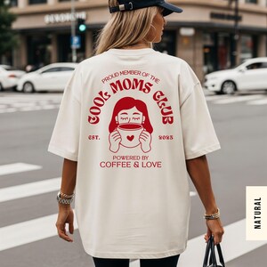 May include: Cream-colored t-shirt with a red graphic that reads "COOL MOMS CLUB" and "POWERED BY COFFEE & LOVE." The design includes an illustration of a woman holding a coffee cup with a heart. The text "EST. 2023" is also included.