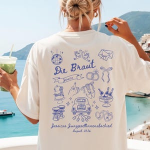 Könnte beinhalten: Ein cremefarbenes T-Shirt mit blauem Grafikdruck auf der Rückseite. Das Design zeigt eine Discokugel, Tauben, Eheringe, eine Torte, eine Hand mit Ring, Schuhe, Champagner, ein Auto mit der Aufschrift 'JUST MARRIED' und 'Die Braut'.