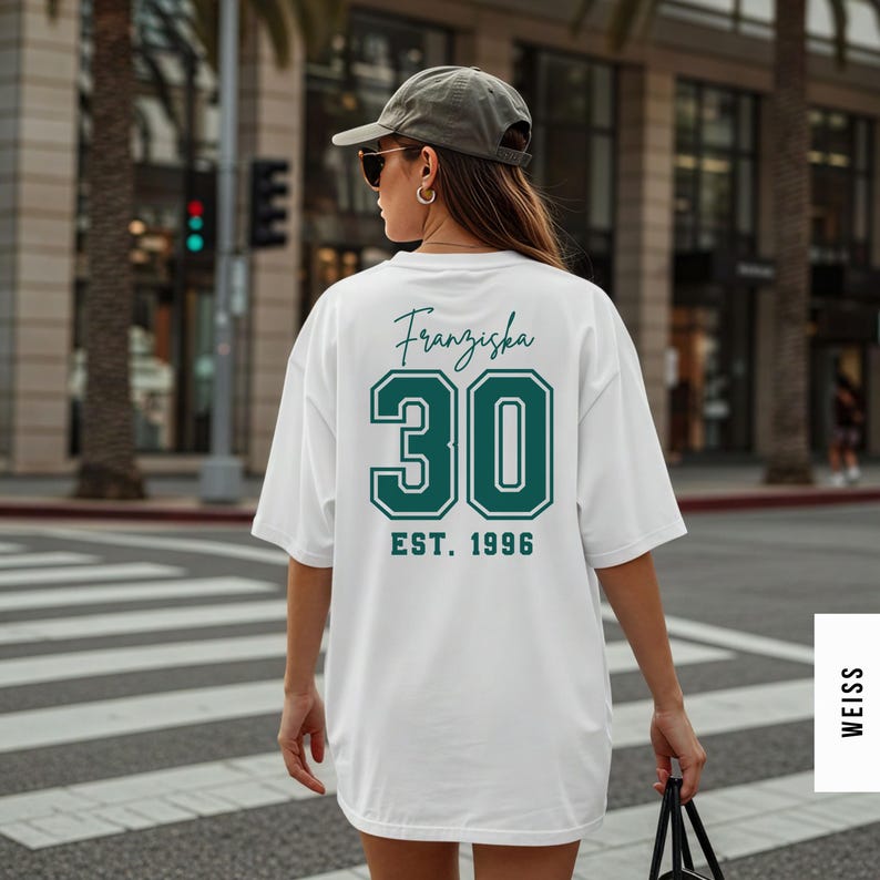 Op de afbeelding: Wit oversized T-shirt met de naam "Franziska", het nummer "30" en "EST. 1996" gedrukt in teal. Het shirt wordt gedragen door een persoon die over een zebrapad loopt.