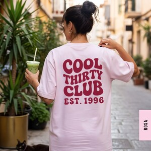 Puede incluir: Una camiseta de manga corta de color rosa claro con texto rojo oscuro en la espalda que dice COOL THIRTY CLUB EST. 1996 en una fuente ondulada y llamativa. La prenda tiene cuello redondo y un corte holgado que resalta el dise&ntilde;o gr&aacute;fico impreso.