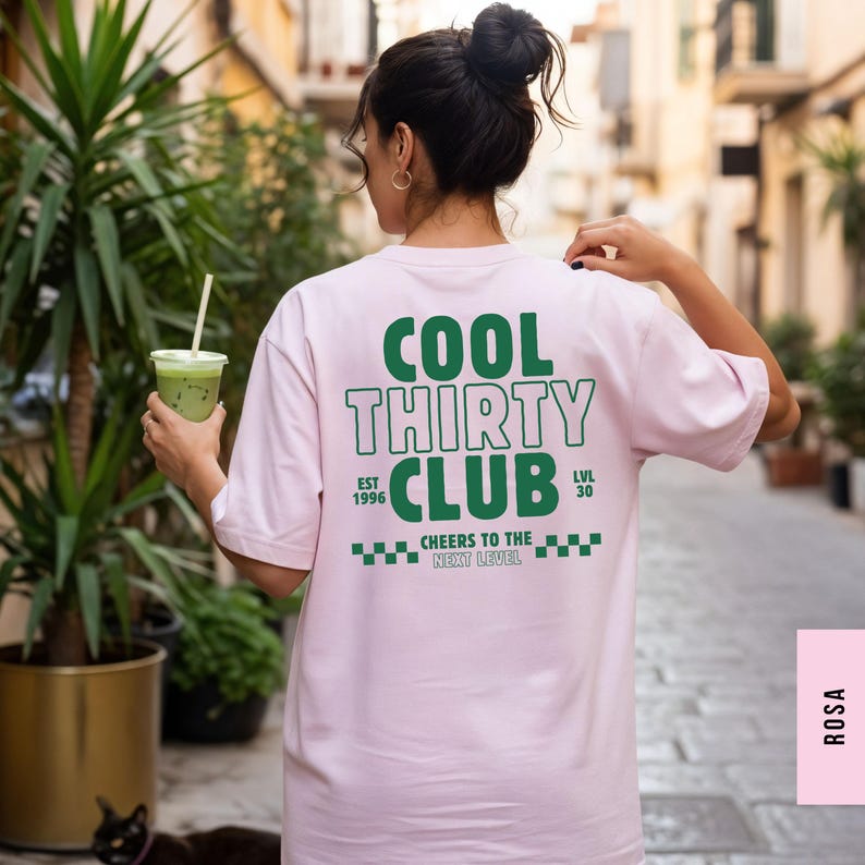 May include: A light pink t-shirt featuring green text on the back that reads COOL THIRTY CLUB, EST 1996, LVL 30, and CHEERS TO THE NEXT LEVEL, accompanied by a green checkered pattern graphic.