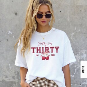 May include: A white short-sleeved t-shirt featuring the text Birthday Girl THIRTY EST. 1996 in red collegiate lettering. Below the text is a graphic of two red cherries tied together with a light pink ribbon bow centered on the chest.