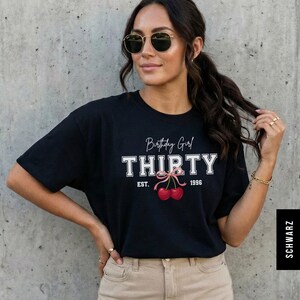 May include: A black short-sleeved t-shirt featuring the white text Birthday Girl THIRTY and EST. 1996. Below the text is a graphic of two red cherries tied together with a light pink ribbon. The shirt has a crew neckline and a standard fit.
