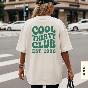 May include: An oversized off-white t-shirt featuring dark green text on the back that reads COOL THIRTY CLUB EST. 1996 in a wavy, retro-inspired font. The shirt has short sleeves and a loose fit, showcasing a clean, solid-colored fabric design.