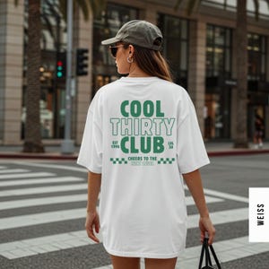 May include: A white oversized t-shirt featuring dark green text on the back that reads COOL THIRTY CLUB, EST 1996, LVL 30, and CHEERS TO THE NEXT LEVEL, accompanied by a checkered pattern. The shirt is worn with a grey baseball cap and sunglasses.