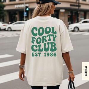 Pu&ograve; includere: Una t-shirt a maniche corte color bianco sporco con testo verde scuro sul retro che recita COOL FORTY CLUB EST. 1986 in un carattere grassetto e ondulato. La maglietta ha una vestibilit&agrave; ampia e rilassata, indossata con un cappellino nero e bracciali dorati.