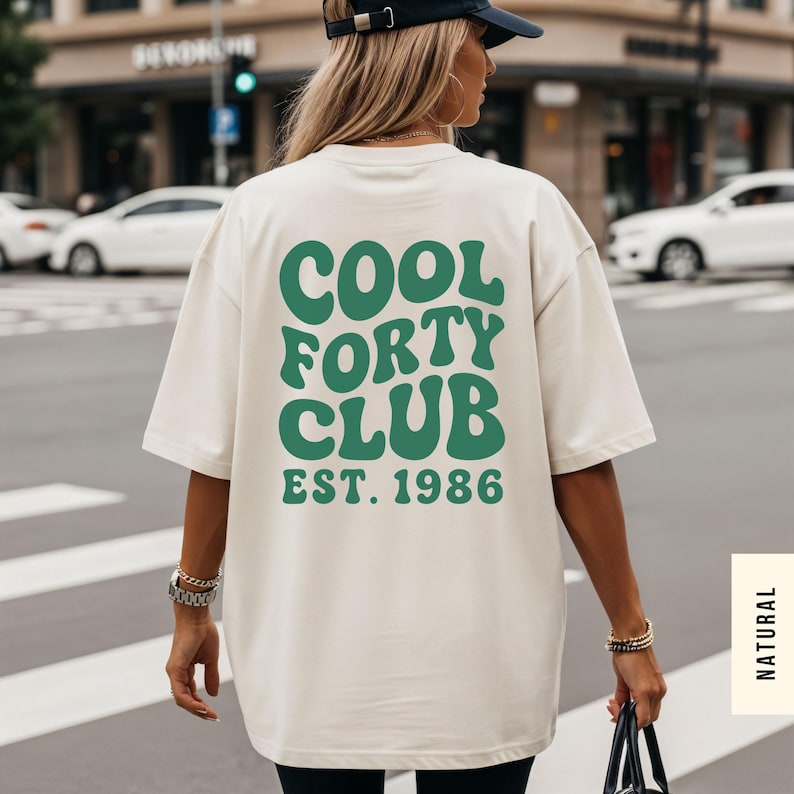 Pu&ograve; includere: Una t-shirt a maniche corte color bianco sporco con testo verde scuro sul retro che recita COOL FORTY CLUB EST. 1986 in un carattere grassetto e ondulato. La maglietta ha una vestibilit&agrave; ampia e rilassata, indossata con un cappellino nero e bracciali dorati.