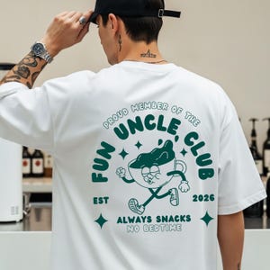 Könnte beinhalten: Weißes T-Shirt mit einem grünen Aufdruck auf dem Rücken. Der Aufdruck zeigt "FUN UNCLE CLUB" mit einer Cartoon-Kaffeetasse. Das Shirt hat auch den Text "EST 2026" und "ALWAYS SNACKS NO BEDTIME".