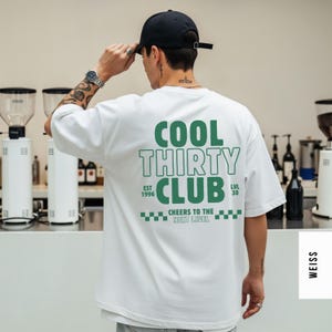 May include: A white short-sleeved t-shirt featuring green text on the back that reads COOL THIRTY CLUB, EST 1996, LVL 30, and CHEERS TO THE NEXT LEVEL, accompanied by a green checkered pattern. The shirt is worn with a black baseball cap.