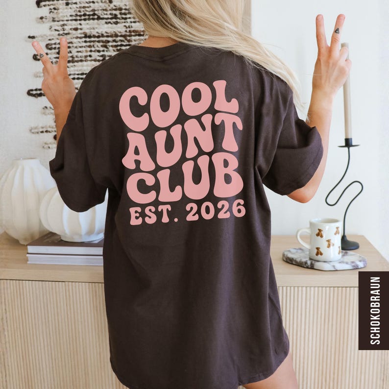Op de afbeelding: Een donkerbruin t-shirt met de roze tekst "COOL AUNT CLUB EST. 2026" in retro-stijl. Het t-shirt wordt gedragen door een persoon met blond haar, die een vredesteken maakt met hun vingers.