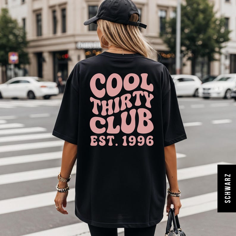 May include: Black t-shirt with the text "COOL THIRTY CLUB EST. 1996" in pink, retro-style lettering. The shirt is worn by a person in an urban setting, with a black cap and accessories.