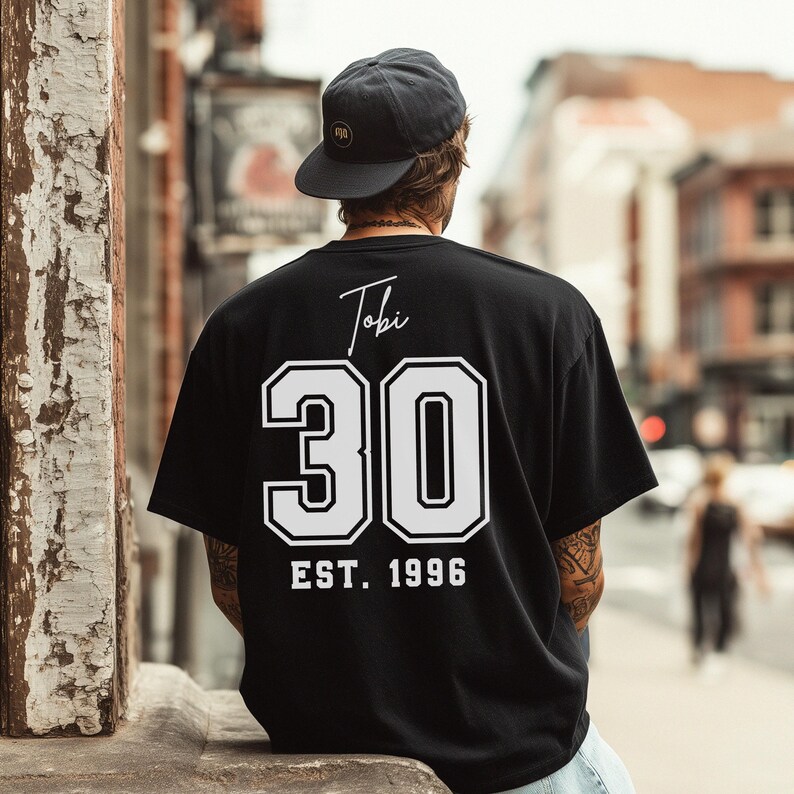 Op de afbeelding: Zwart T-shirt met de naam "Tobi" in script boven het nummer "30" in witte blokletters. Onder het nummer staat "EST. 1996" in wit. De persoon op de afbeelding draagt een zwarte pet.