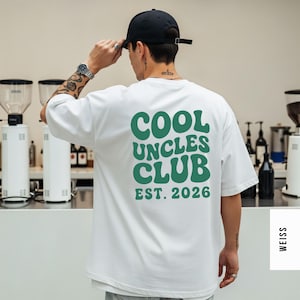 Könnte beinhalten: Weißes T-Shirt mit dem grünen Text "COOL UNCLES CLUB EST. 2026", eine schwarze Baseballkappe und eine silberne Uhr. Das Shirt hat eine lockere, übergroße Passform.