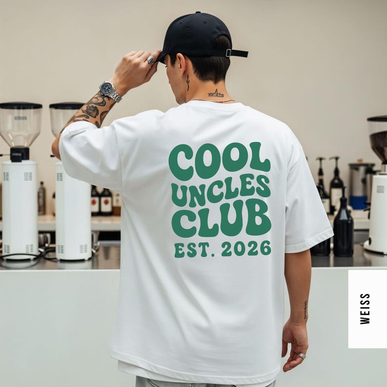 K&ouml;nnte beinhalten: Wei&szlig;es T-Shirt mit dem gr&uuml;nen Text "COOL UNCLES CLUB EST. 2026", eine schwarze Baseballkappe und eine silberne Uhr. Das Shirt hat eine lockere, &uuml;bergro&szlig;e Passform.