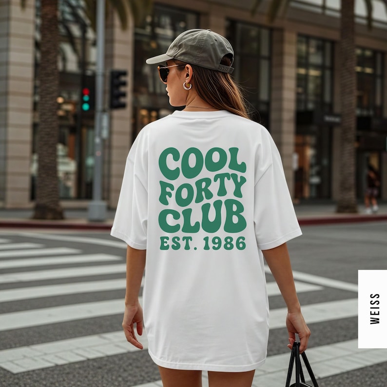 Pu&ograve; includere: Una t-shirt bianca con la scritta verde scuro COOL FORTY CLUB EST. 1986 in un carattere ondulato di ispirazione retr&ograve; sul retro. La maglia ha maniche corte e una vestibilit&agrave; ampia. &Egrave; indossata insieme a un berretto da baseball grigio.