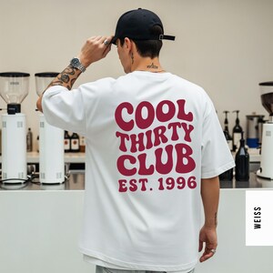 Puede incluir: Una camiseta blanca de manga corta con el texto granate COOL THIRTY CLUB EST. 1996 impreso en la espalda con una fuente ondulada y negrita. La camiseta tiene un corte holgado y relajado, y se lleva con una gorra negra y un reloj de pulsera plateado.