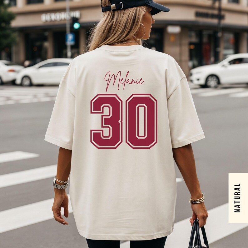 May include: An oversized cream-colored t-shirt featuring the name Melanie in a dark red script font above the large dark red number 30 in a collegiate block style. The shirt has short sleeves and a loose fit, displayed from the back view.