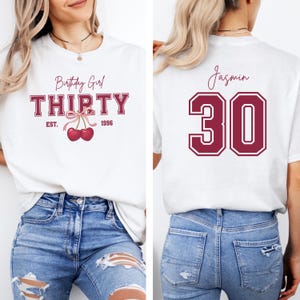 May include: A white t-shirt featuring a front graphic with the text Birthday Girl THIRTY EST. 1996 and a pair of red cherries with a pink bow. The back of the shirt displays the name Jasmin above the large maroon number 30 in a collegiate font.