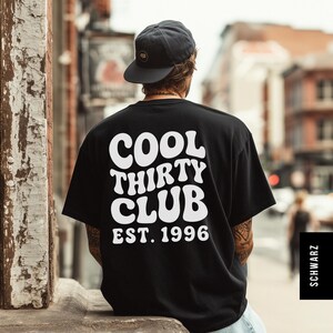 May include: A black short-sleeve t-shirt featuring white text on the back that reads COOL THIRTY CLUB EST. 1996 in a wavy font. The shirt is paired with a dark gray baseball cap with a small circular logo on the front panel.