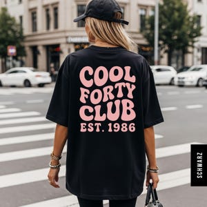 May include: Black t-shirt with the text "COOL FORTY CLUB EST. 1986" in pink, a design for a 40th birthday. The shirt is worn by a person walking on a crosswalk in a city.