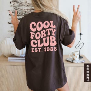 May include: A dark brown t-shirt with the text "COOL FORTY CLUB EST. 1986" in pink, displayed on the back. The shirt is a relaxed fit, and the design has a retro, groovy aesthetic.