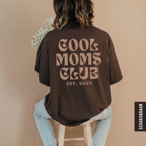 Könnte beinhalten: Ein dunkelbraunes T-Shirt mit kurzen Ärmeln, das auf der Rückseite den Text COOL MOMS CLUB in einer großen, wellenförmigen, hellbraunen Schrift zeigt. Darunter befindet sich der Text EST. 2023 in derselben Farbe. Das Shirt wird zu einer hellblauen Jeans getragen.