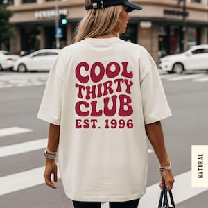 Puede incluir: Una camiseta de corte oversize en color crema con texto rojo oscuro en la espalda que dice COOL THIRTY CLUB EST. 1996 en una fuente ondulada de estilo retro. La camiseta tiene mangas cortas y cuello redondo para un ajuste relajado.