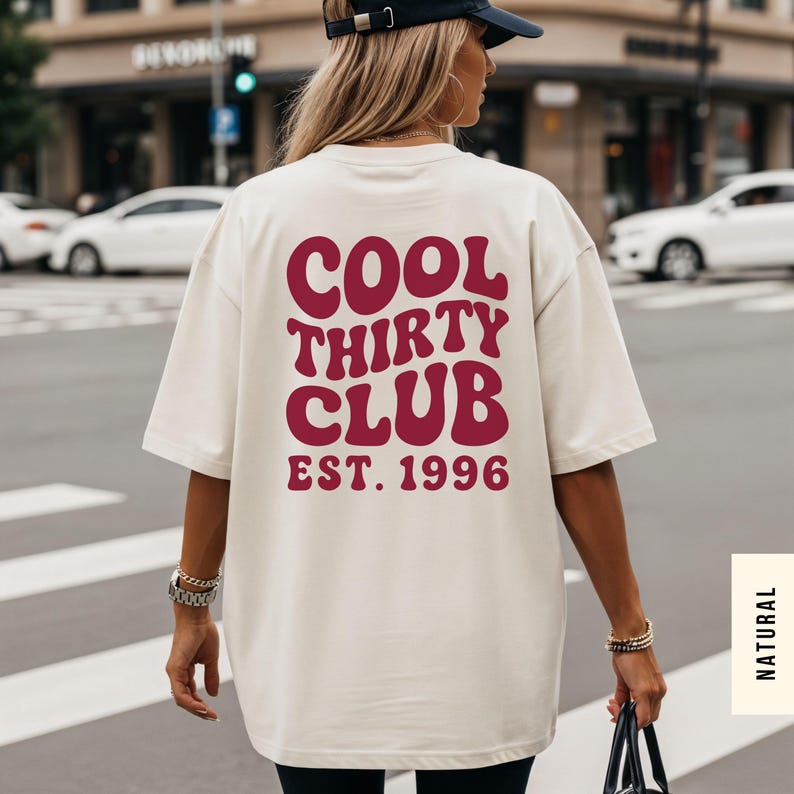 Puede incluir: Una camiseta de corte oversize en color crema con texto rojo oscuro en la espalda que dice COOL THIRTY CLUB EST. 1996 en una fuente ondulada de estilo retro. La camiseta tiene mangas cortas y cuello redondo para un ajuste relajado.