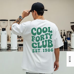 Pu&ograve; includere: Una t-shirt bianca a maniche corte con una grande scritta verde ondulata sul retro che recita COOL FORTY CLUB EST. 1986. La maglia ha una vestibilit&agrave; ampia e rilassata con girocollo ed &egrave; indossata con un cappellino da baseball nero tinta unita.