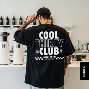 May include: A black short-sleeved t-shirt featuring white text on the back that reads COOL THIRTY CLUB, EST 1996, LVL 30, and CHEERS TO THE NEXT LEVEL, accompanied by a checkered pattern graphic. The shirt is worn with a black baseball cap.