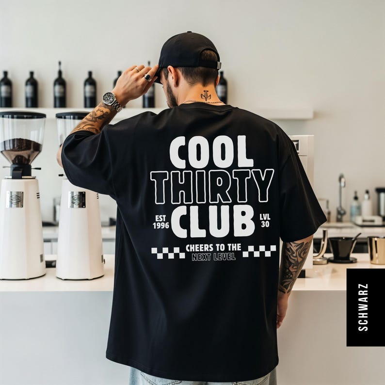 May include: A black short-sleeved t-shirt featuring white text on the back that reads COOL THIRTY CLUB, EST 1996, LVL 30, and CHEERS TO THE NEXT LEVEL, accompanied by a checkered pattern graphic. The shirt is worn with a black baseball cap.