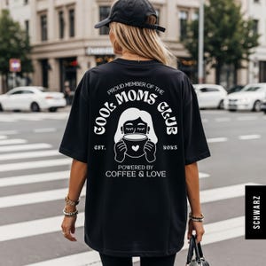 May include: Black oversized t-shirt with a white graphic on the back that reads "Proud Member of the Cool Moms Club" with a coffee cup illustration. The text "Powered by Coffee & Love" is below the graphic. The shirt is worn by a person.