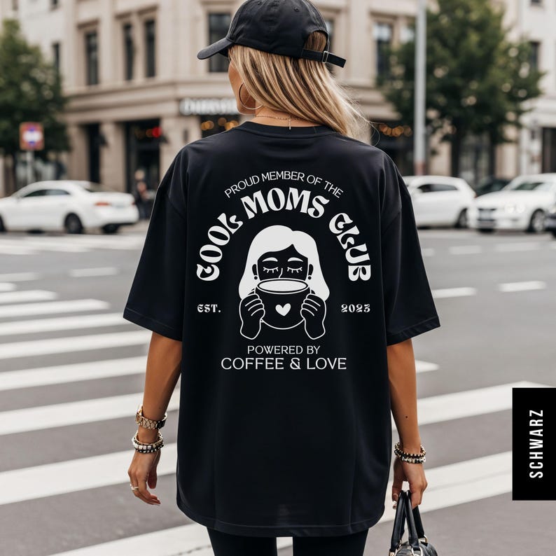May include: Black oversized t-shirt with a white graphic on the back that reads "Proud Member of the Cool Moms Club" with a coffee cup illustration. The text "Powered by Coffee & Love" is below the graphic. The shirt is worn by a person.