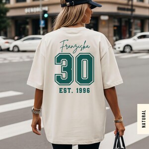 Op de afbeelding: Cr&egrave;mekleurig T-shirt met de naam "Franziska", het nummer "30" en "EST. 1996" op de rug gedrukt in teal. Het shirt is oversized en heeft korte mouwen. De persoon draagt een zwarte baseball cap.