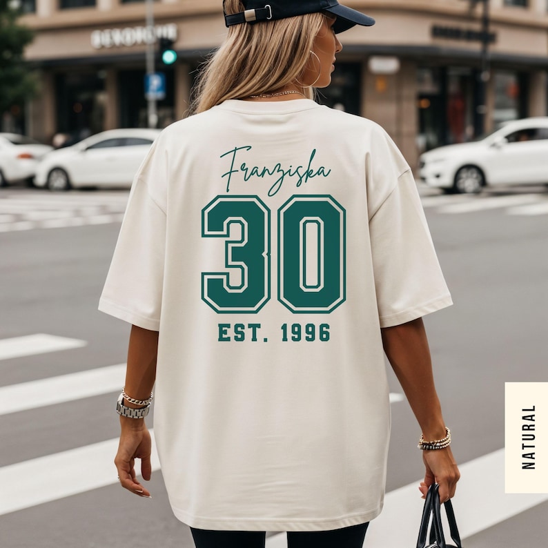 Op de afbeelding: Cr&egrave;mekleurig T-shirt met de naam "Franziska", het nummer "30" en "EST. 1996" op de rug gedrukt in teal. Het shirt is oversized en heeft korte mouwen. De persoon draagt een zwarte baseball cap.