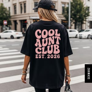 Op de afbeelding: Zwart T-shirt met de roze tekst "COOL AUNT CLUB EST. 2026" in een retro lettertype. De persoon draagt het shirt en loopt op een zebrapad in een stedelijke omgeving.