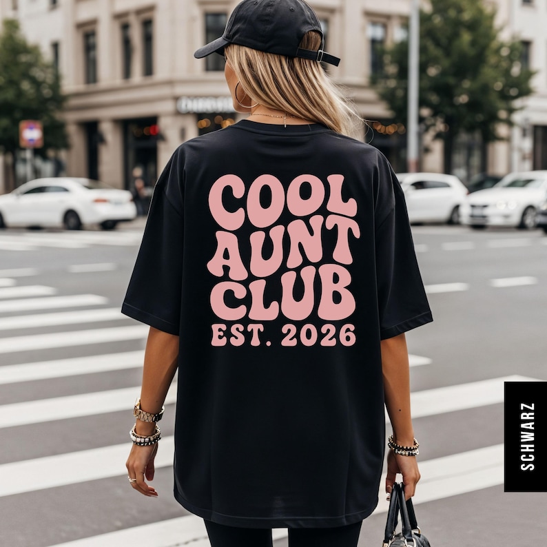 Op de afbeelding: Zwart T-shirt met de roze tekst "COOL AUNT CLUB EST. 2026" in een retro lettertype. De persoon draagt het shirt en loopt op een zebrapad in een stedelijke omgeving.