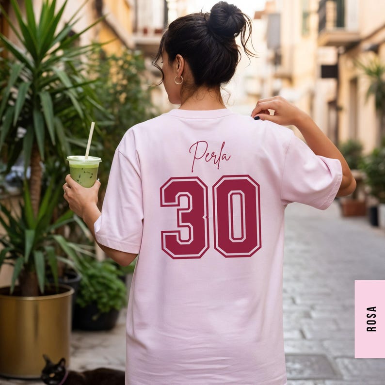 May include: A light pink short-sleeved t-shirt featuring the name Perla in a dark red script font above the large dark red number 30 with a white outline printed on the back. The shirt has a crew neckline and a relaxed fit design.