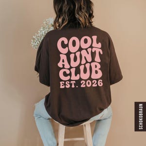 Op de afbeelding: Een donkerbruin t-shirt met de roze tekst "COOL AUNT CLUB EST. 2026" in een retro lettertype. De persoon draagt het t-shirt en zit op een houten kruk. De tekst is vetgedrukt.