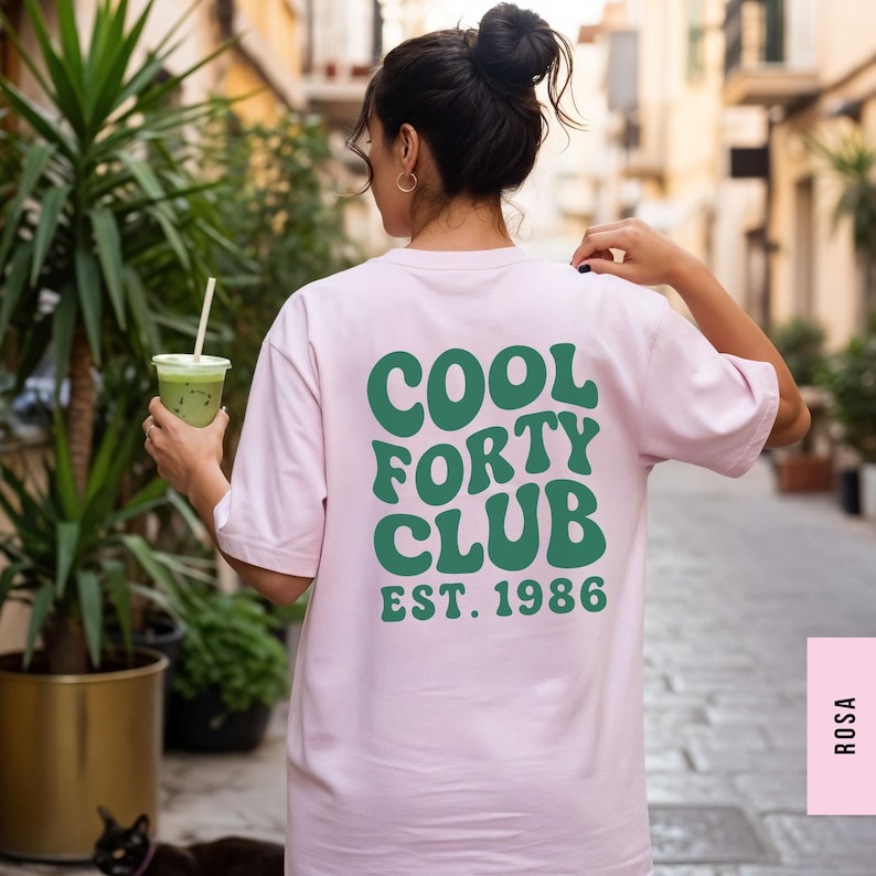 Pu&ograve; includere: Una t-shirt a maniche corte color rosa chiaro con testo verde scuro sul retro che recita COOL FORTY CLUB EST. 1986 in un carattere audace e ondulato. La maglia presenta un girocollo e una vestibilit&agrave; rilassata con la grafica ben visibile.
